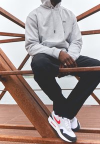 Harmaa puuvillane huppari, kus on tasku, mustad püksid ja valged tossud, millel on punased ja hallid aktsendid. Istub metallistruktuuri peal.