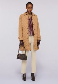 Hellbrauner gesteppter Mantel, dunkelrotes Lederoberteil, cremefarbene Hose, dunkle Handschuhe und Handtasche. Das Model trägt elegante schwarze Stiefeletten.