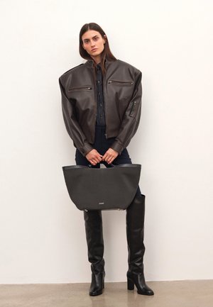 Kvinde iført en oversized mørk læderjakke, sorte høje støvler, holder en stor sort totebag, stående mod en ensfarvet hvid væg.