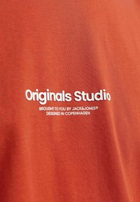Rød bomulds T-shirt med hvid tekst: "Originals Studio," "Bragt til dig af JACK&JONES," og "Designet i København." Blød tekstur.