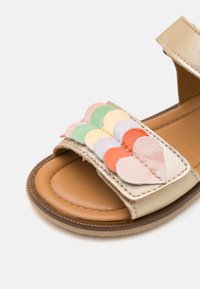 Guld syntetiske sandaler har en tan sål og en bred rem dekoreret med overlappende, multifarvede lædderflager i pastelnuancer.