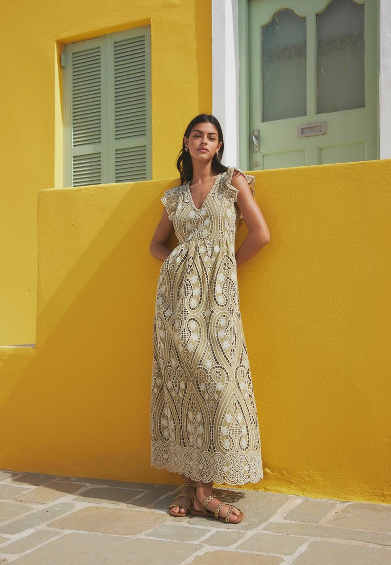 Robe maxi en beige clair avec un motif en dentelle paisley blanche complexe et des manches volantes, associée à des sandales à lanières marron devant un mur jaune.