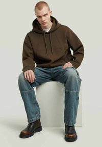 Bruine hoodie met een frontzak, in combinatie met blauwe denim cargo broeken met patchworkdetails en zwarte laarzen met bruine accenten.