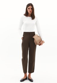 Donna in una maglietta bianca a maniche lunghe e pantaloni marroni a vita alta, che tiene in mano una pochette beige mentre con l'altra mano si infila in tasca, su uno sfondo bianco.