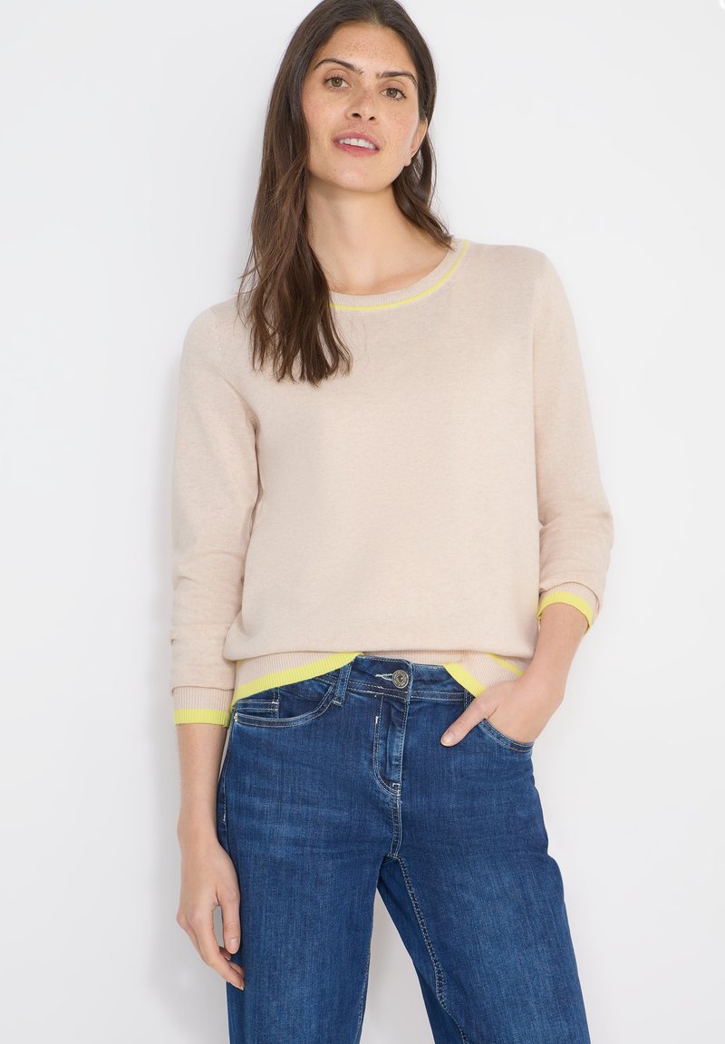 Pull beige clair avec des accents jaune vif au niveau du col et des poignets, porté avec un jean en denim bleu. Présente un col rond et une coupe décontractée.