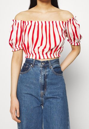 Kvinde iført hvid off-shoulder crop top med røde striber og højtaljede blå denimjeans, hånd på hoften mod ensartet baggrund.