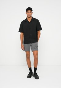 Schwarzes, kurzärmeliges Hemd mit Knöpfen, kombiniert mit grauen Shorts. Das Model trägt schwarze Schuhe und Socken. Glatter Stoff und lässige Passform.