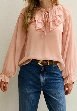 Femme portant un chemisier rose transparent à volants avec des manches longues et un jean bleu, accessoirisée d'une ceinture marron avec une boucle dorée.