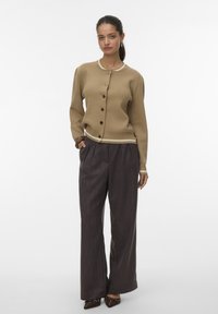 Vrouw staand, gekleed in een tan kleurige, met knopen gesloten cardigan, losse donkere gestreepte broek en spitse bruine schoenen, tegen een effen witte achtergrond.