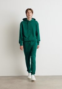 Grønn hettejakke og matchende joggebukser i mykt stoff, med snor i midjen, frontlomme og hvite sneakers. Minimalistisk design.