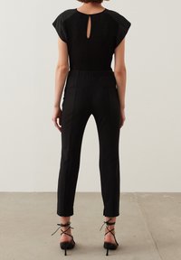 Combinaison noire avec un haut côtelé, des manches courtes et un détail en trou de serrure au dos. Pantalons fuselés avec deux poches arrières. Portée avec des talons à lanières.