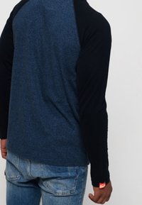 Man in een blauw en zwart langslievend shirt met lichtblauwe spijkerbroek, staande met zijn rug naar de camera.