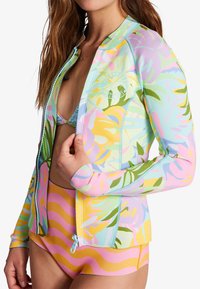 Camisa de protección multicolor con patrones de hojas tropicales en tonos pastel, con cierre de cremallera y mangas largas. Usada sobre un bikini.