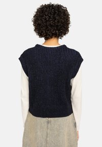 Gestructureerde marineblauwe gebreide vest met korte mouwen, ribbelontwerp en een cropped lengte, gelaagd over een licht lange mouwtop.