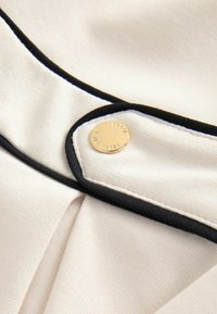 Tessuto crema con dettagli in piping nero, caratterizzato da un bottone dorato con il nome del brand inciso. La superficie è liscia e leggermente strutturata.