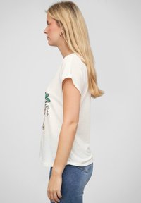 T-shirt blanc à manches courtes avec un imprimé graphique floral coloré. Tissu doux, coupe décontractée. Associé à un jean bleu, vu de côté.