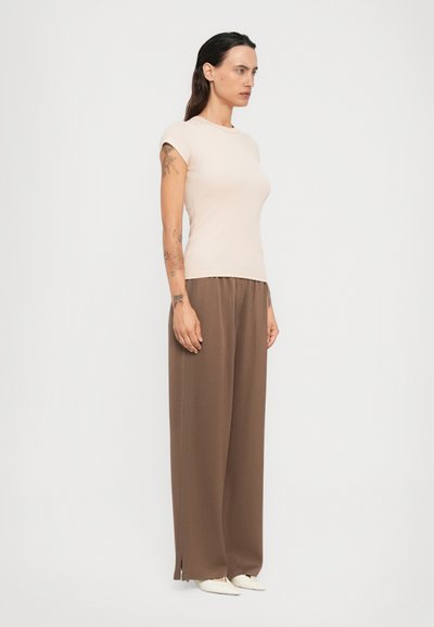 Donna con lunghi capelli scuri e tatuaggi, che indossa un top beige a maniche corte, pantaloni larghi marroni e scarpe bianche a punta, in piedi davanti a uno sfondo bianco.