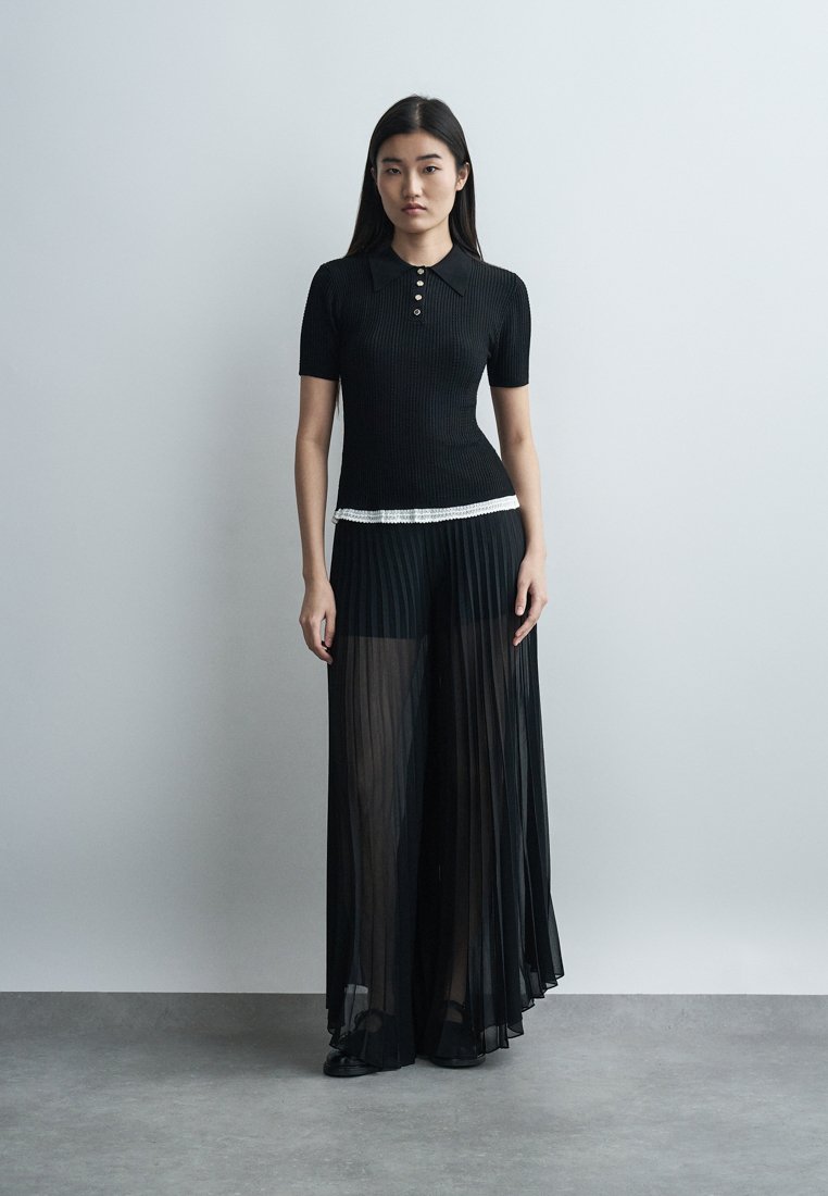 Femme portant un polo noir à manches courtes et un pantalon large noir plissé transparent, debout contre un mur gris clair uni.