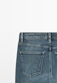 Achteraanzicht van een blauwe spijkerbroek met twee achterzakken, riemlussen en zichtbare stiksels op een witte achtergrond.