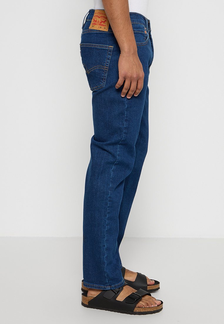 Blaue Jeans aus Denim mit geradem Schnitt, Gürtelschlaufen und einem braunen Lederpatch auf der Rückseite. Getragen mit schwarzen Sandalen auf einem weißen Hintergrund.