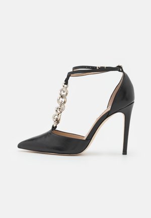 Zapato negro de piel con punta afilada y tacón alto, detalle de cadena plateada y cierre de hebilla en la correa del tobillo, mostrado sobre fondo blanco.
