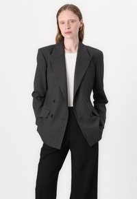 Dobbeltspent blazer i mørkegrått med hvite striper, med hakkede slag og strukturerte skuldre. Kombinert med en hvit skjorte og svarte bukser.