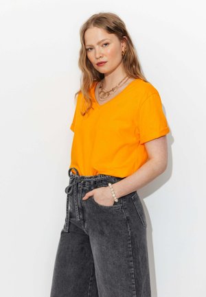 Femme aux longs cheveux ondulés, portant un haut orange vif à manches courtes et un jean noir taille haute, main dans la poche, portant des bijoux en or et un bracelet de perles.