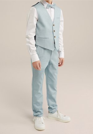 Chłopiec ubrany w jasnoniebieską kamizelkę, pasujące spodnie, białą koszulę, muszkę oraz białe trampki, stojący na jednolitym tle.