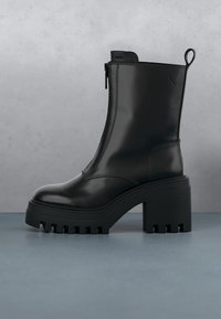 Bottine en cuir noir avec semelle crantée épaisse, talon bloc, fermeture éclair frontale et languette de tirage sur une surface et un fond gris.