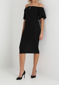 Robe noire décolletée épaules avec manches courtes à volants, silhouette ajustée et ourlet à hauteur du genou. Portée avec des talons hauts noirs.