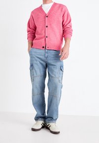 Muž v růžovém zapínacím kardiganu, bílém tričku, světle modrých cargo džínách a bílých teniskách s hnědými pruhy stojí před bílým pozadím.