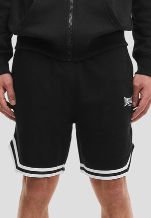 Uomo che indossa pantaloncini sportivi neri con bordo bianco e logo Lonsdale London sulla coscia sinistra, abbinati a una felpa nera con zip.