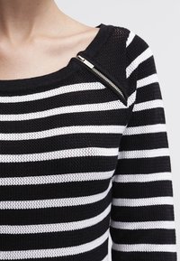 Gestreifter Pullover in Schwarz und Weiß, mit strukturiertem Strick, weitem Ausschnitt und seitlichem Reißverschlussdetail an der linken Schulter.