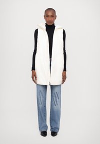 Padded white vest with a high collar and zip closure, worn over a black turtleneck, paired with blue wide-leg jeans featuring a logo print.