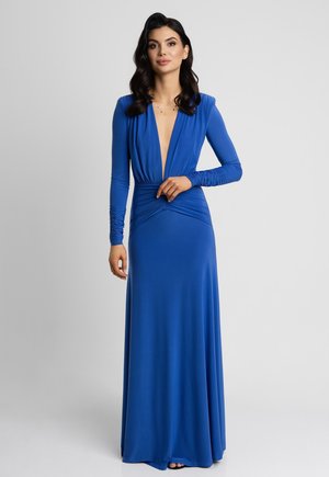 Kobieta z ciemnymi włosami, ubrana w długą, niebieską suknię z długim rękawem, sięgającą do podłogi, z głębokim dekoltem w serek i marszczonym pasem, stojąca na tle jednolitego szarego tła.