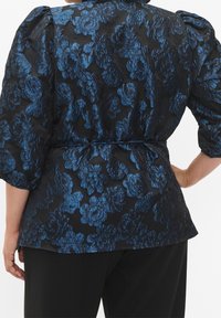 Donna che indossa un blusa nera con ricami floreali blu e maniche a sbuffo, abbinata a pantaloni neri, vista da dietro.