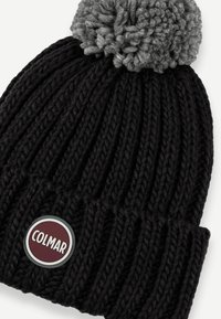 Juodas megztas beanie kepurė su pilku pom-pomu viršuje. Yra raukšlėta tekstūra ir apvalus raudonas etiketė su baltu "COLMAR" prekės ženklu.