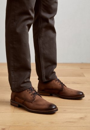 Zapatos marrones de cuero con textura y cordones, usados con pantalones marrón oscuro, de pie sobre un suelo de madera clara.
