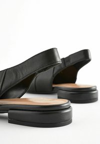 Fekete bőr, hátul nyitott cipők kerek orral, lapos talppal és textúrázott pánttal. A cipők sima bélésű talppal rendelkeznek.