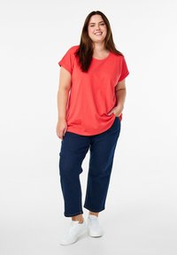 Koraalkleurig T-shirt met korte, opgerolde mouwen, ontspannen pasvorm, gecombineerd met donkerblauwe denim broek en witte sneakers. Soepele stoftextuur.