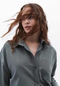 Camicia grigia con bottoni realizzata in un tessuto liscio. Presenta un colletto a punta, maniche lunghe e una tasca applicata sul lato sinistro.