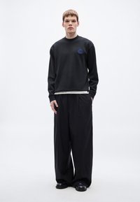 Μαύρο φούτερ με στρογγυλή λαιμόκοψη και μπλε κεντημένο ετικέτα, συνδυασμένο με φαρδιά μαύρη παντελόνια, φορεμένο με μαύρα παπούτσια. Απλός σχεδιασμός.