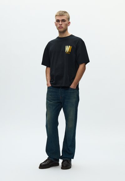 Μαύρο βαμβακερό T-shirt με χρυσό λογότυπο 'WN' στο αριστερό στήθος, συνδυασμένο με μπλε τζιν παντελόνι και μαύρα παπούτσια.