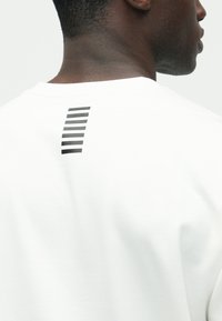 Tricou alb din bumbac, cu un imprimeu grafic cu dungi verticale negre pe partea superioară a spatelui, mâneci scurte și decolteu rotund.