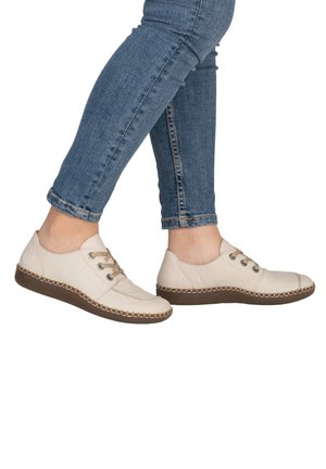 Beige leather shoes with brown rubber soles, featuring laces and stitched detailing on the toe and around the sole edge. Worn with blue jeans.