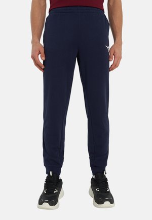Uomo che indossa pantaloni da jogging aderenti blu navy con fascia elastica in vita e scarpe sportive nere, in piedi contro uno sfondo semplice.