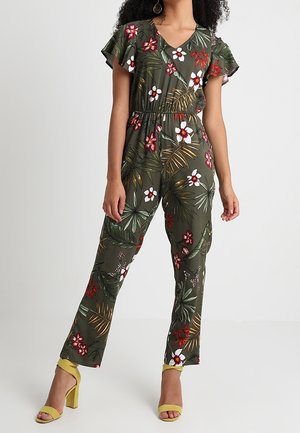 Combinaison fleurie vert foncé avec manches courtes à volants, col en V, taille élastiquée, et sandales à talons jaunes. Motifs floraux et feuilles remarquables.