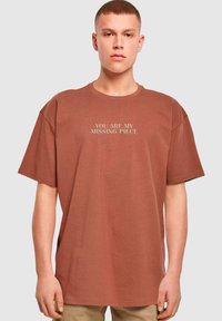 Brun oversized t-shirt lavet af bomuld, med korte ærmer og trykt tekst "DU ER MIN MANGELDEL" i en lysere farve på fronten.