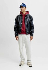 Zwarte leren bomberjack, bordeauxrode hoodie, grijze joggingbroek en beige sneakers. Het model draagt een blauwe pet met een logo.