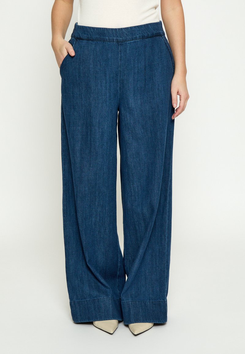 Frau trägt locker sitzende, weit geschnittene dunkelblaue Jeans mit Seitentaschen und weißen spitzen Schuhen, steht vor einem schlichten Hintergrund.